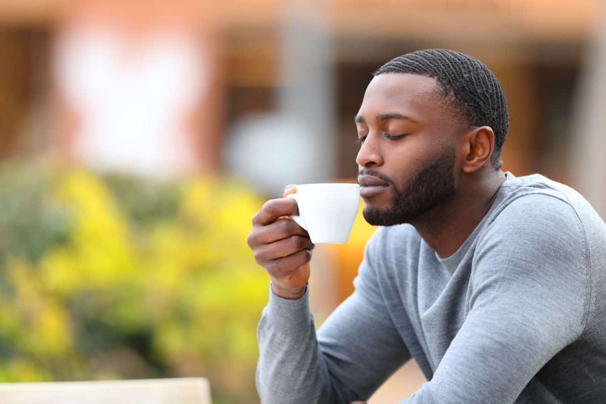 Man smelling a cup of coffee in an outdoor setting containing a bush of yellow flowers and a blurry storefront