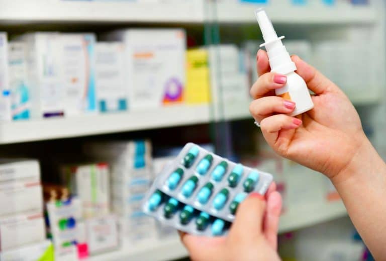 A blurry background of a drugstore shelf. Two hands are in frame and in focus. One hand holds two packets of green and blue pills while the other hand holds a nasal spray bottle.