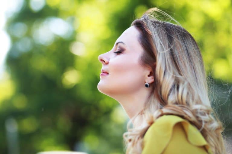 Woman in profile in an outdoor setting. She is facing the left side of the frame with her eyes closed and her chin tilted slightly up. She has a content expression. The background is heavily blurred tree canopy with enough light to indicate it is not a forest. 